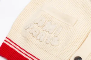 Amiri unisex woven ADC stripes cardigan featuring a cream-colored body, red striped accents, and a textured "AMI PARIS" logo on the pocket.