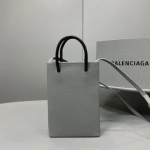 Mini Grey Balenciaga crossbody bag made of luxurious calf leather, featuring iconic logo, stylish design, and black handles, displayed on a table with a branded box in the background.