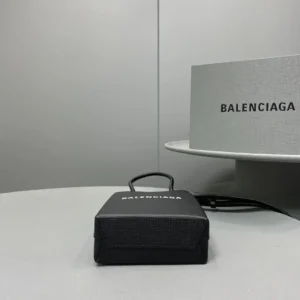 Mini black Balenciaga crossbody bag displayed on a gray surface next to a Balenciaga box, showcasing its trendy design and luxury branding.