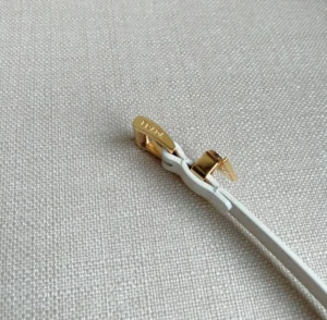 Elegant PRADA Saffia belt detail featuring a soft triangular metal logo decoration and white strap against a textured background.