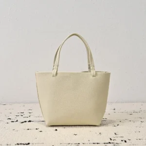 The Row Park tote bag in cream color, featuring minimalist design, soft lychee patterned cowhide material, and dual handles, displayed on a textured surface.