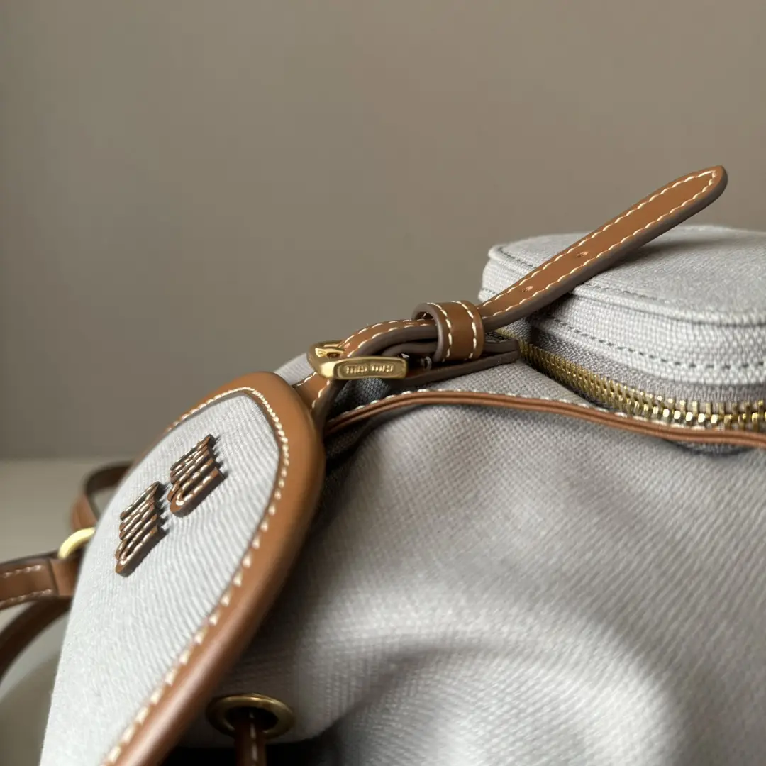 Close-up of MIU New College Style Backpack showing leather strap, zipper detail, and signature logo on gray canvas material.