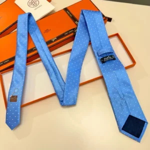 Hermès Letter Men's Tie in light blue with polka dots, displayed on a white surface alongside an orange Hermès box, emphasizing luxury fashion accessories.