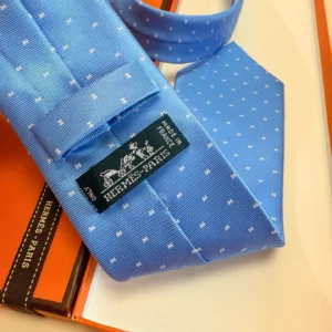 Hermès Letter Men's Tie in blue with white polka dots and logo tag, displayed on an orange box, showcasing premium fashion accessory.