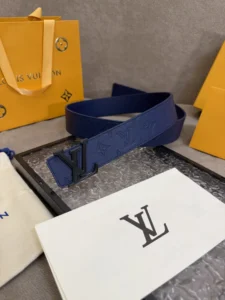 LV Shadow belt with Monogram Shadow displayed on a gray surface, accompanied by Louis Vuitton packaging and branding materials, showcasing luxury accessory details.