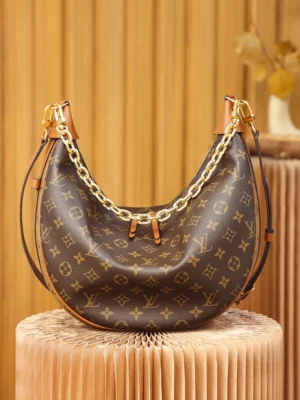 Montaigne BB Monogram canvas handbag by Louis Vuitton, featuring a crescent shape, gold chain detailing, and iconic monogram pattern, displayed on a textured pedestal.