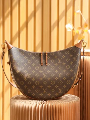 Montaigne BB Monogram canvas handbag by Louis Vuitton, featuring a crescent shape and brown monogram pattern, displayed against a warm, textured background.