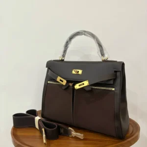 Hermes Kelly Lakis 28 bag in ebony and gold, featuring dual zippers and a detachable strap, displayed on a wooden surface.