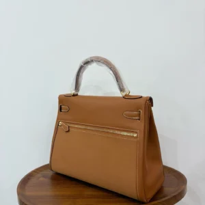 Hermes Kelly Lakis 28 handbag in gold brown leather with gold accents, displayed on a wooden table.