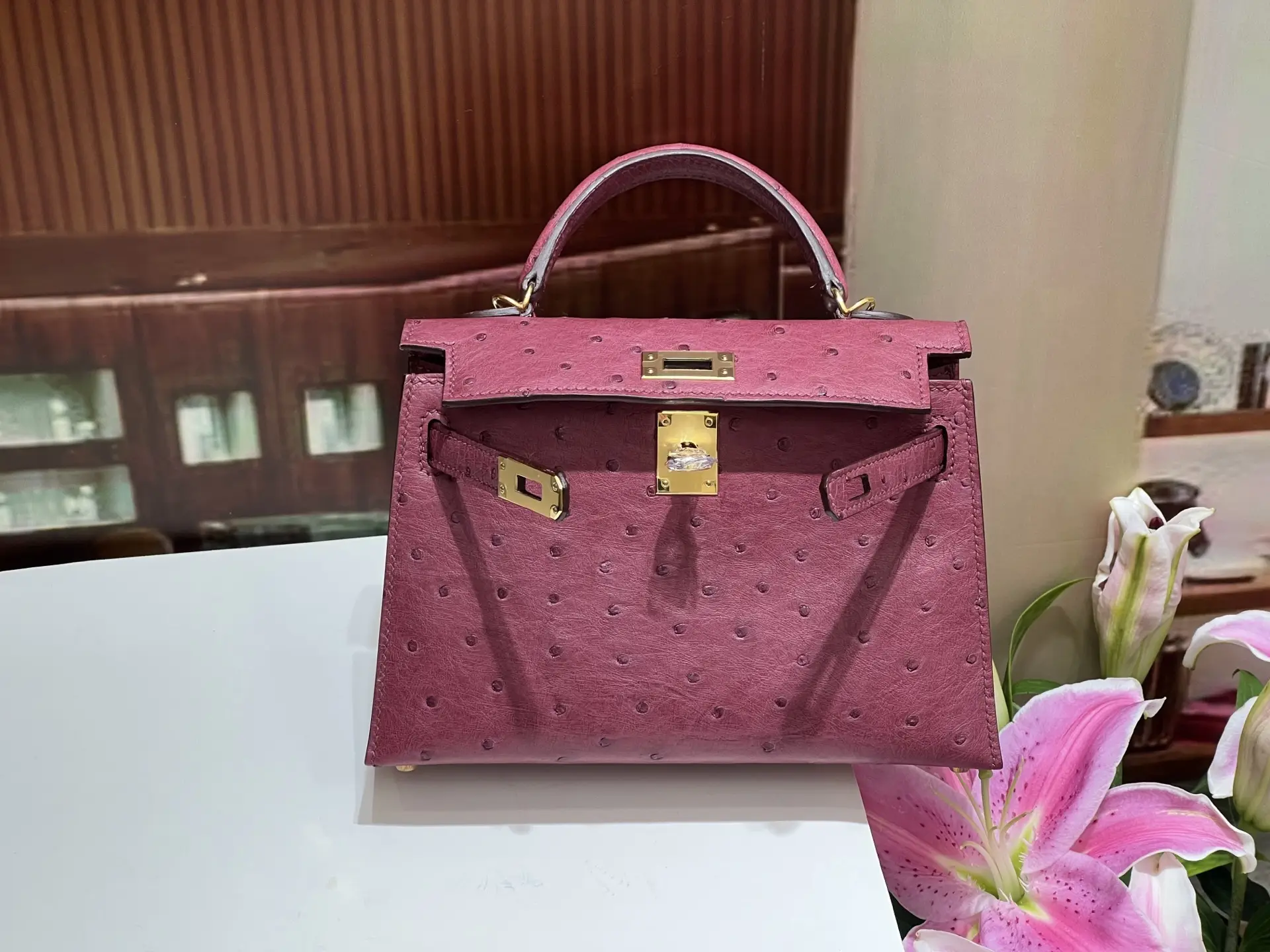 Kelly's second-generation 19CM wine red gold buckle handbag made from South African imported KK ostrich skin, featuring a luxurious design and high-quality craftsmanship, displayed alongside pink lilies.