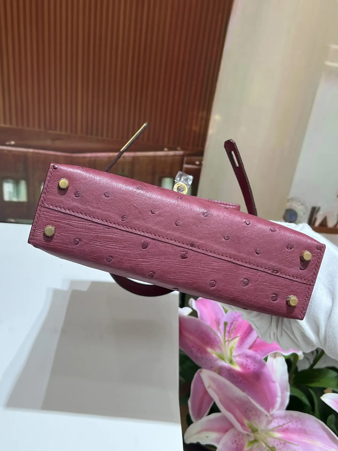 Wine red KK ostrich skin handbag with gold buckle, showcasing high-quality craftsmanship, photographed in a floral setting.