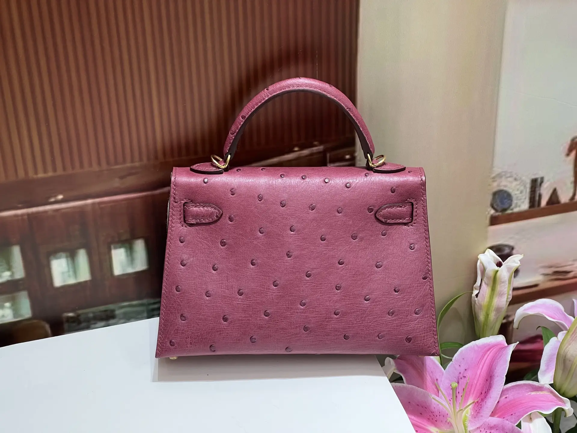 Kelly's second-generation 19CM wine red gold buckle handbag made from South African imported KK ostrich skin, featuring a structured design and textured leather, displayed alongside pink lilies.