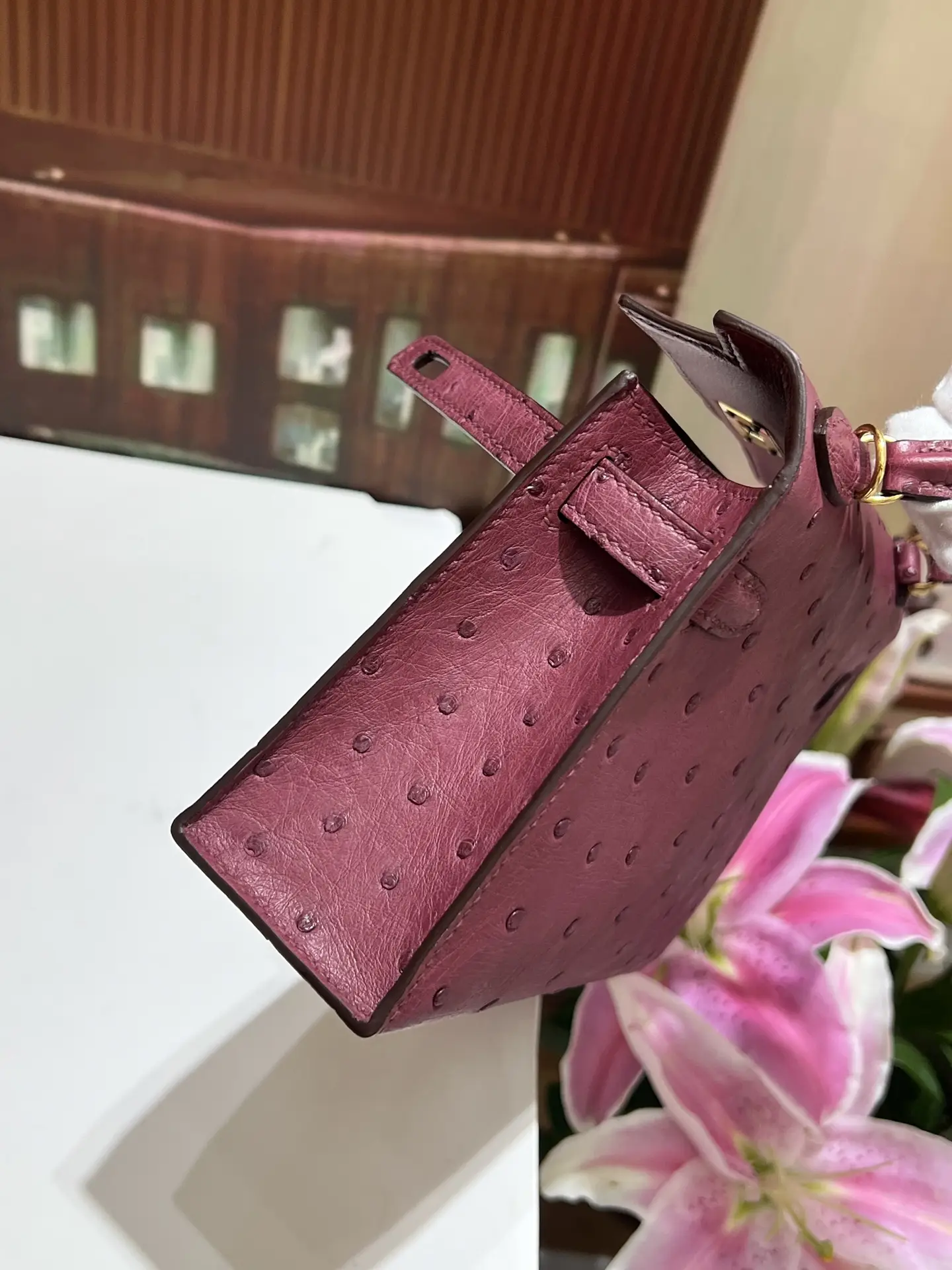 Wine red Kelly handbag with gold buckle, crafted from South African imported KK ostrich skin, displayed beside pink lilies.