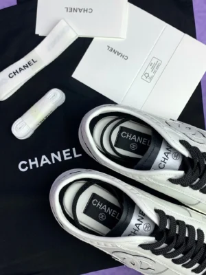 Chanel 25P series casual sports shoes in pure white with black laces, displayed on a branded Chanel dust bag and accompanied by care instructions and extra laces.