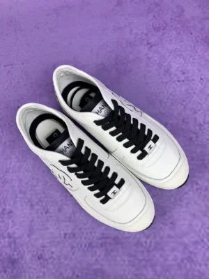 Chanel 25P series casual sports shoes in pure white with black laces and logo, displayed on a purple background.