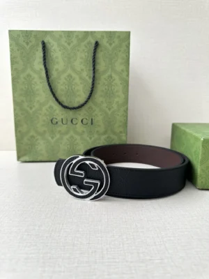 Gucci belt featuring interlocking double G buckle, displayed with signature green Gucci shopping bag and box, showcasing cowhide craftsmanship in black and grey color options.
