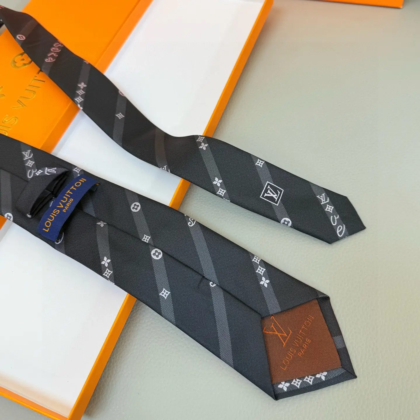 Silk LV tie featuring diagonal stripes, LV logo, and Monogram flowers, displayed on an orange Louis Vuitton box.