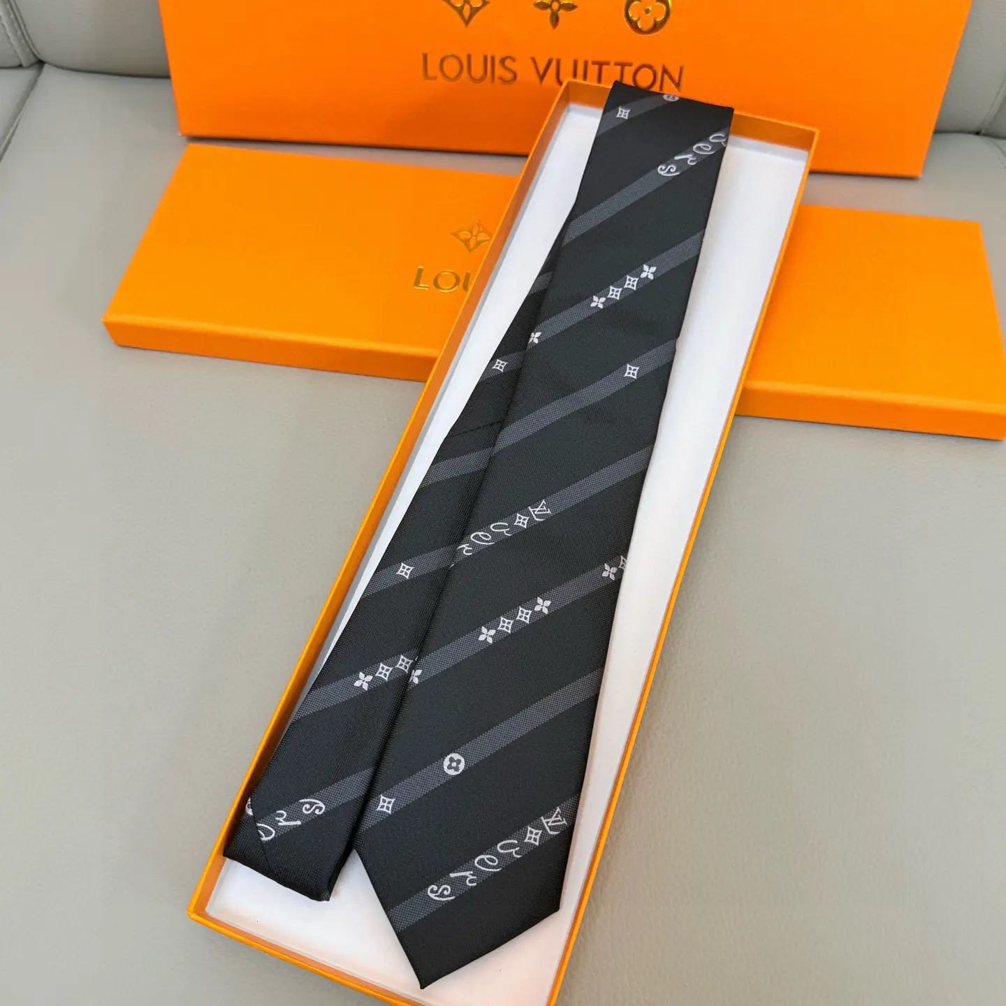 Silk tie featuring LVVers logo and Monogram flowers in diagonal stripe design, displayed in an orange Louis Vuitton box.