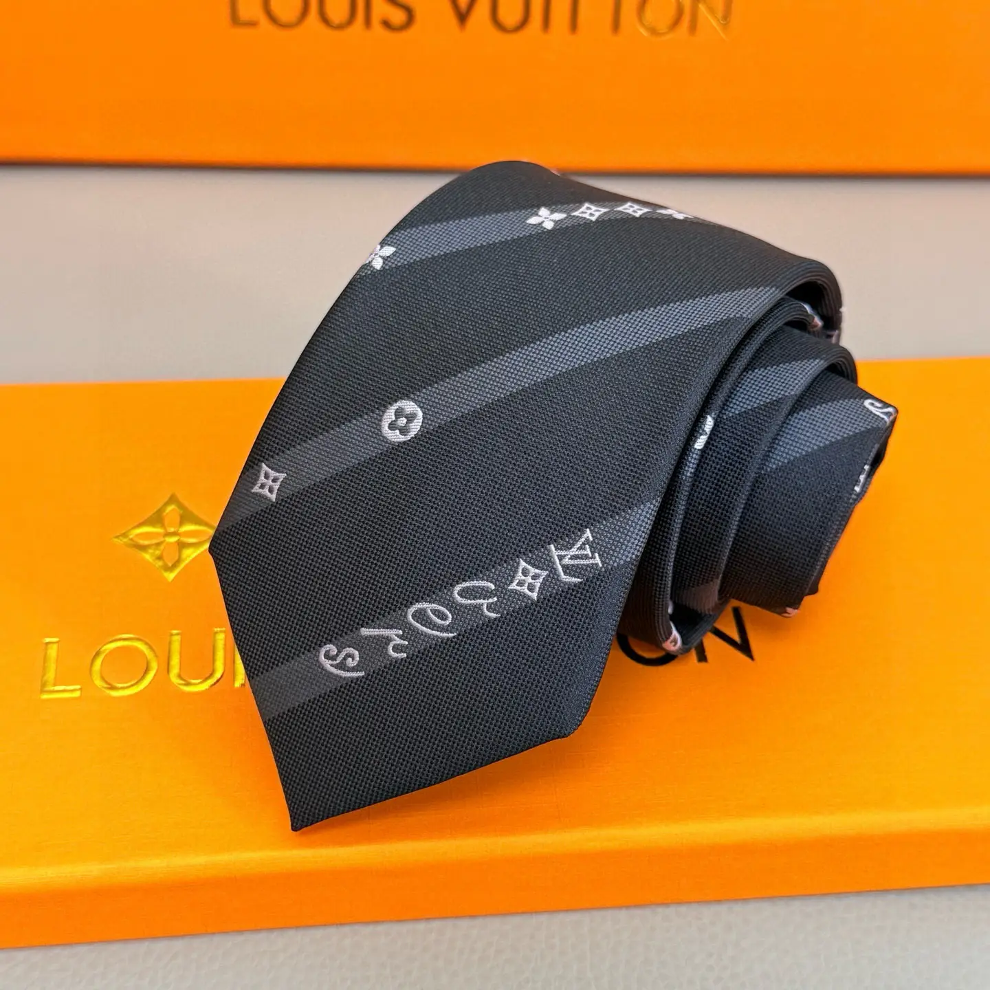 Silk tie featuring LVVers logo and Monogram flowers on diagonal stripes, placed on an orange Louis Vuitton box, showcasing luxury accessory design.