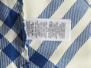 The image shows a fabric care and composition label sewn onto a blue and white patterned textile, with information printed in multiple languages. There is no product brand, model, or specific product type visible to generate the requested SEO-optimized alt text according to your rules. Please provide an image with a clear view of the product showcasing brand, model, and key features for the correct alt text generation.
