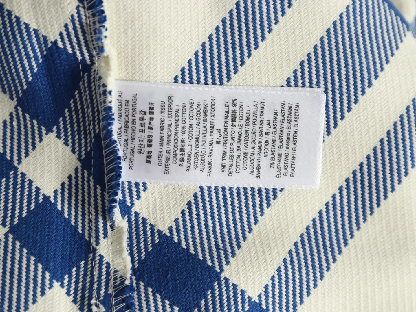 The image shows a fabric care and composition label sewn onto a blue and white patterned textile, with information printed in multiple languages. There is no product brand, model, or specific product type visible to generate the requested SEO-optimized alt text according to your rules. Please provide an image with a clear view of the product showcasing brand, model, and key features for the correct alt text generation.