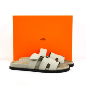 Hermes flat one-word slippers in light gray, displayed against an orange box, showcasing luxury footwear design and branding.