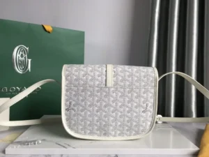 Goyard Belv é d è re Single Mailman Bag in light gray with adjustable shoulder strap, featuring minimalist design and embossed logo, displayed on a marble surface alongside a green Goyard shopping bag.