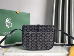 Goyard Belv é d è re Single Mailman Bag displayed on a surface with a green Goyard shopping bag in the background, showcasing its elegant design, adjustable shoulder strap, and signature pattern.