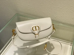 Dior Bobby East-West Bag in white leather with gold hardware, showcasing a sleek crescent shape and detachable shoulder strap, displayed in a glass case.