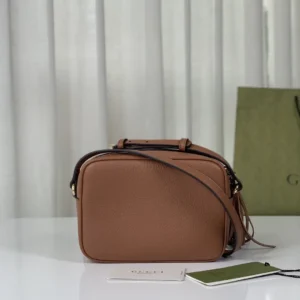 Apricot leather tote bag displayed on a table, featuring a shoulder strap, brand tag, and a Gucci shopping bag in the background.