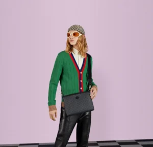 Model wearing a green cardigan with red and blue accents, black leather pants, and sunglasses, holding a black GG Supreme canvas Interlocking Double G Postman Bag against a pastel pink background.