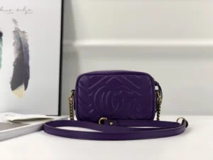 Purple Gucci GG Marmont small shoulder bag with interlocking G logo, featuring a gold chain strap, placed beside decorative feathers and a magazine.