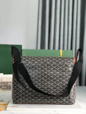 Goyard Citadin Mailman Bag in grey and black with adjustable shoulder strap, displayed against a backdrop of green boxes and soft drapery, highlighting its stylish design and functionality as a versatile messenger bag.