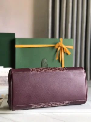 Goyard Saigon Tote bag in burgundy leather with signature design, displayed on a marble surface with a green gift box and yellow ribbon in the background.