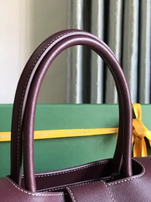Goyard Saigon Tote bag handles in rich burgundy leather, showcasing elegant stitching and design, with a green background and yellow ribbon, emphasizing luxury and style.