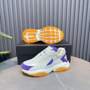 AMIRI Trainer replica sneaker in white, purple, and orange, displayed on a surface with a black box and green plant in the background.