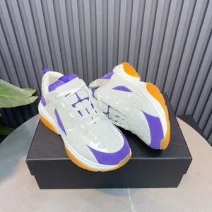 AMIRI Trainer replica sneakers in white, purple, and orange colorway, displayed on a black shoebox with a textured background.