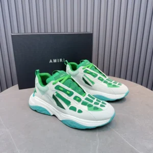 Amiri Trainer sneakers in white and green with unique design elements, displayed on a gray surface next to an Amiri branded box.