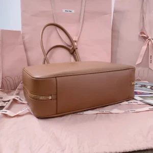 Elegant Miu Miu bowling handbag in smooth tan leather, featuring a zippered compartment, displayed on a soft pink background with matching bags, highlighting its sophisticated multifunctional design.