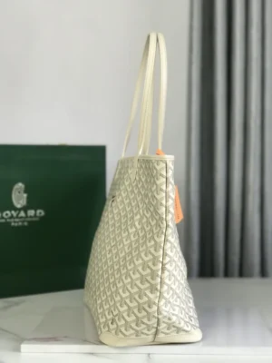 Goyard White Tote Bag displayed on a marble surface, showcasing its textured design, cream color, and elegant straps, with a green Goyard box in the background.
