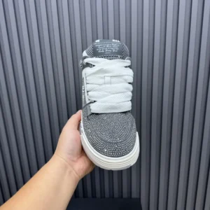 AMIRI Trainer sneaker held in hand, featuring a textured black and white design with prominent laces, showcased against a striped background, emphasizing luxury footwear.