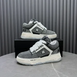 AMIRI Trainer sneakers in black and white, featuring a textured upper, prominent logo, and modern design, displayed on a gray surface against a striped background.