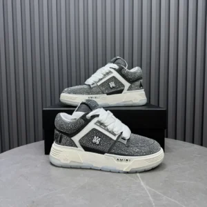 AMIRI Trainer sneakers in gray and white, featuring a textured upper and logo, displayed on a black box against a striped background.