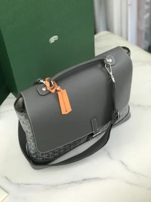 Goyard Citadin Mailman Bag in gray leather and canvas, featuring a detachable shoulder strap and a stylish tag, positioned on a marble surface with a green box in the background.