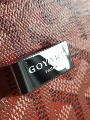 Goyard Paris logo tag on a textured red and black background, highlighting luxury branding associated with the Goyard Belvedere II Bag.