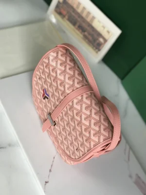 Metallic leather crossbody shoulder purse in pink, featuring Goyard's signature design and adjustable strap, showcased on a marble surface with a green background.