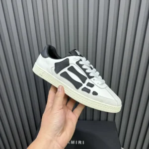 AMIRI Trainer sneakers in white and black, showcasing a modern design with unique paneling, held against a textured gray background.