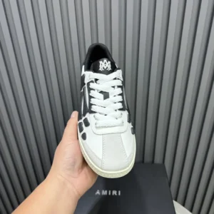 AMIRI Trainer sneaker held in hand, featuring a black and white design, displayed on a branded box with vertical gray stripes in the background.