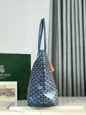 Goyard Pearl Blue tote bag displayed from the side, showcasing its distinctive pattern and leather material, with a green Goyard shopping bag in the background.