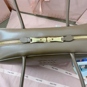 Handmade spliced leather briefcase by MIU MIU, featuring a smooth, soft exterior with a gold zipper closure, set against a light background with MIU MIU branding.